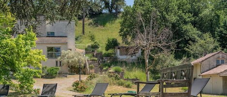 A pool area with lounge chairs, a building with a tiled roof, and a hillside with trees.