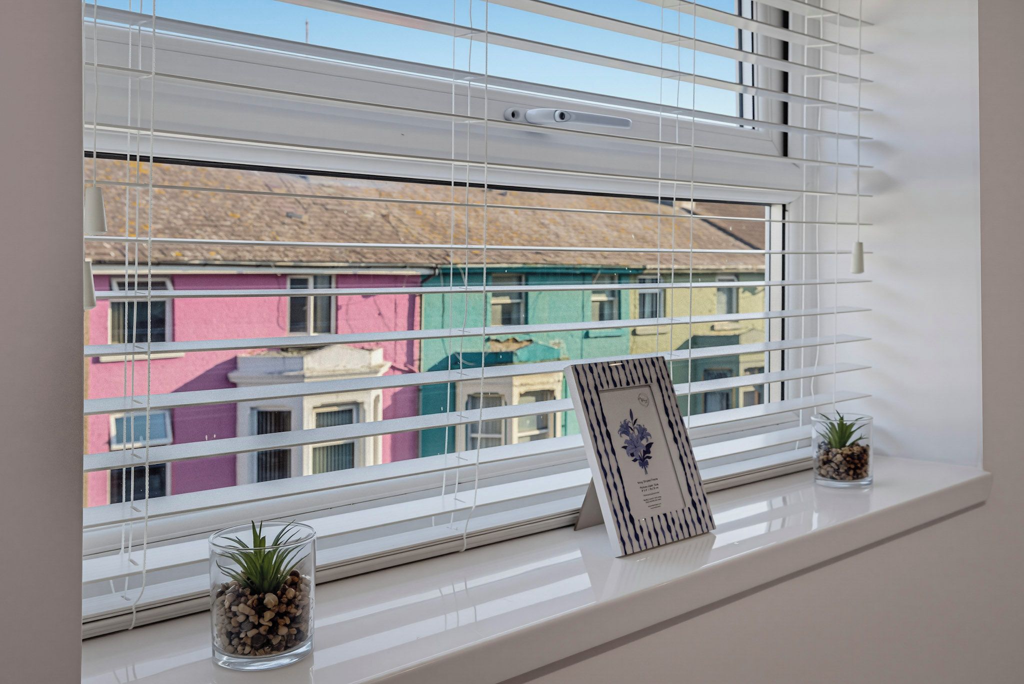 Una ventana con persianas horizontales, un cuadro enmarco y dos pequeñas macetas con plantas en el alféizar.