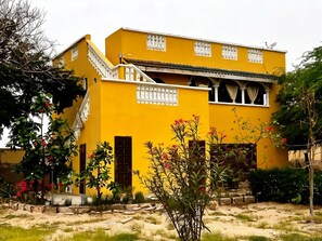 A two-story building with a yellow facade, a balcony with white railings, and a garden with various plants and flowers.