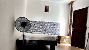 Ein Schlafzimmer mit einem Bett, einem Standventilator, einem gemusterten Kopfteil, einem Holzschrank und einer Tür.