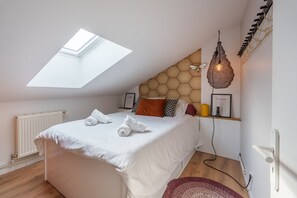 Une chambre dotée d’une lucarne, d’un lit recouvert de draps blancs, d’un luminaire suspendu et d’un mur orné d’un motif géométrique.