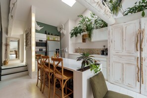 Une cuisine moderne avec des meubles blancs, un coin repas avec des chaises en bois et une grande plante en pot.
