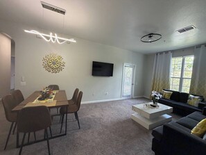 A modern living room with a dining area, a flat-screen TV mounted on the wall, a sofa set, and a decorative wall piece. - Beautiful Apartment near Disney (Kissimmee)