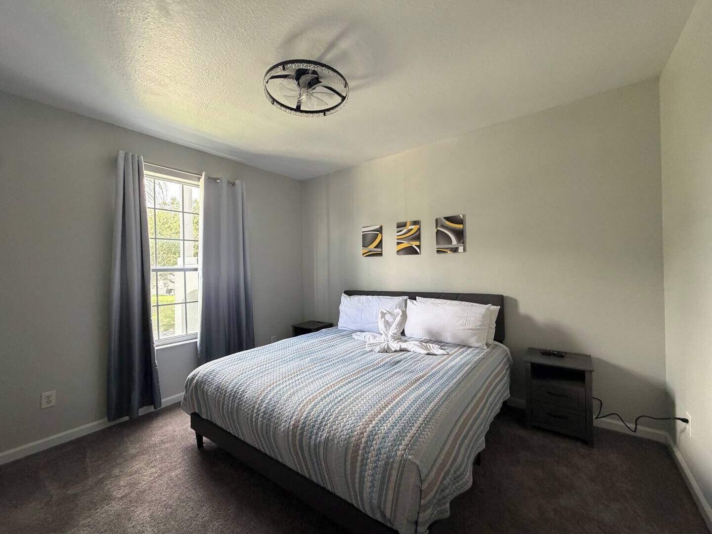 Un dormitorio con cama, mesita de noche, ventilador de techo y ventana con cortinas.