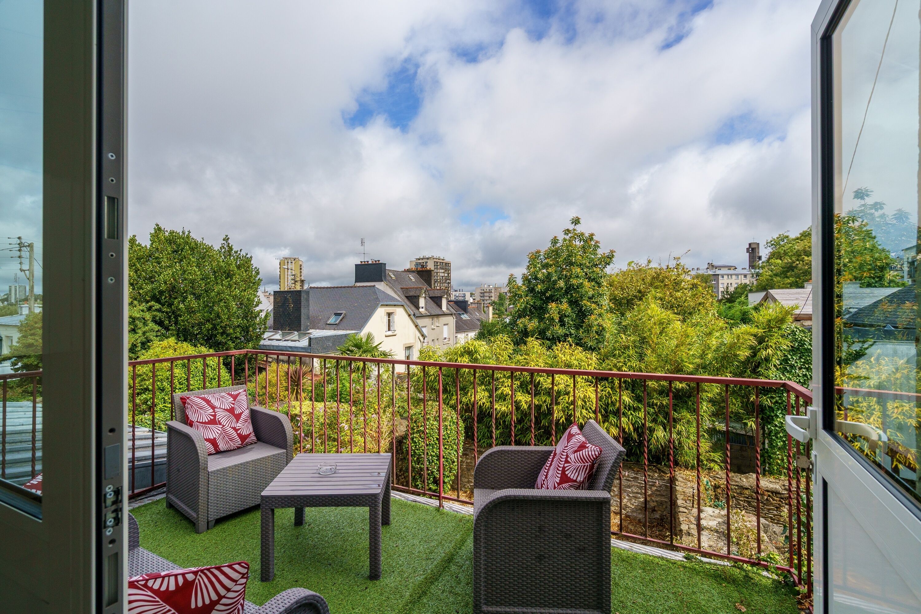 A balcony with wicker furniture, a table, and a view of surrounding buildings and trees.