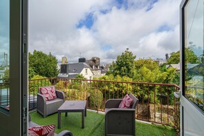 Un balcon agrémenté de meubles en osier, d’une table et offrant une vue sur les bâtiments et les arbres environnants.