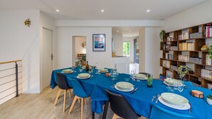 Uma sala de jantar com uma mesa comprida posta para seis pessoas, rodeada por cadeiras, e uma estante de livros repleta à direita.
