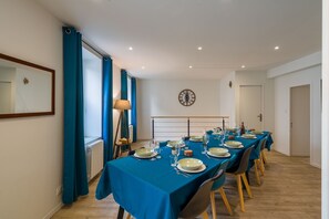 Un coin repas avec une longue table dressée pour le repas, des rideaux bleus, une horloge au mur et un radiateur.