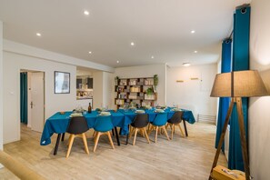 Un coin repas avec une longue table dressée pour six personnes, entourée de chaises, avec une bibliothèque et une lampe en arrière-plan.