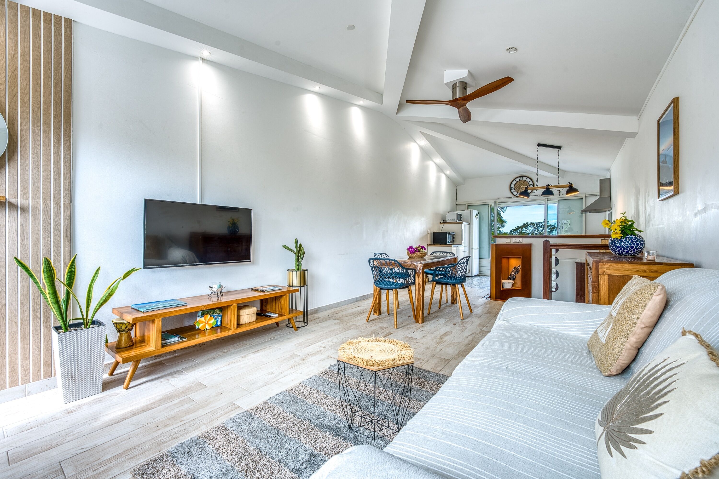 Ein modernes Wohnzimmer mit einem Flachbildfernseher, Holzmöbeln, einem Essbereich mit Stühlen und einem Deckenventilator.