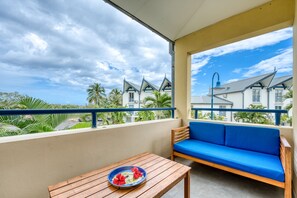 Ein Balkon mit einem Holztisch und einer blauen Couch, mit Blick auf ein Wohngebiet mit Häusern und Palmen.