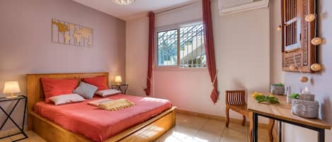 A bedroom with a wooden bed, a bedside table with a lamp, a window with curtains, and a wall-mounted air conditioner.