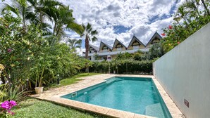 Uma piscina com borda de pedra, rodeada por vegetação exuberante e um edifício com estilo arquitetônico marcante ao fundo.