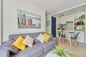 A modern living room with a grey sofa, yellow and white patterned cushions, a dining area with a white table and chairs, and a kitchen in the background. - Cocon Entre Mer, Nature et Histoire (Blonville-sur-Mer)