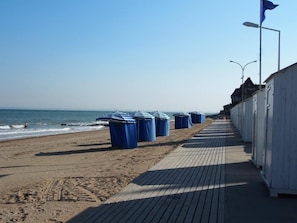 A beachside walkway with a wooden deck, a row of blue trash bins, and a white building with a flag. - Cocon Entre Mer, Nature et Histoire (Blonville-sur-Mer)