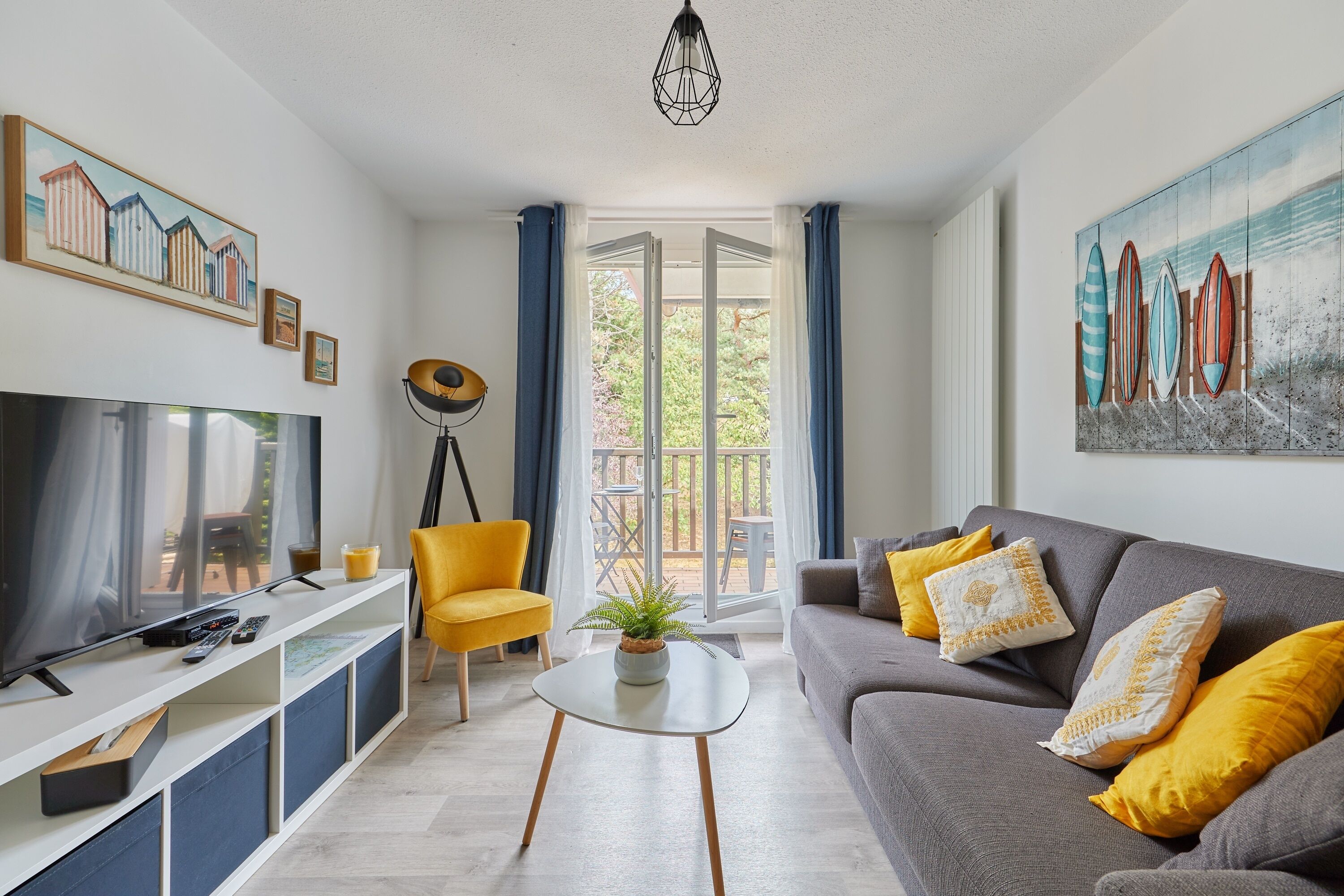 Un salon moderne avec un téléviseur à écran plat, un canapé avec des coussins jaunes, une table basse et un tableau représentant des planches de surf.
