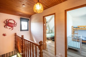 Eine Holztreppe mit Geländer, eine Holzbalkendecke und im angrenzenden Raum ein Bett mit weißem Gestell.