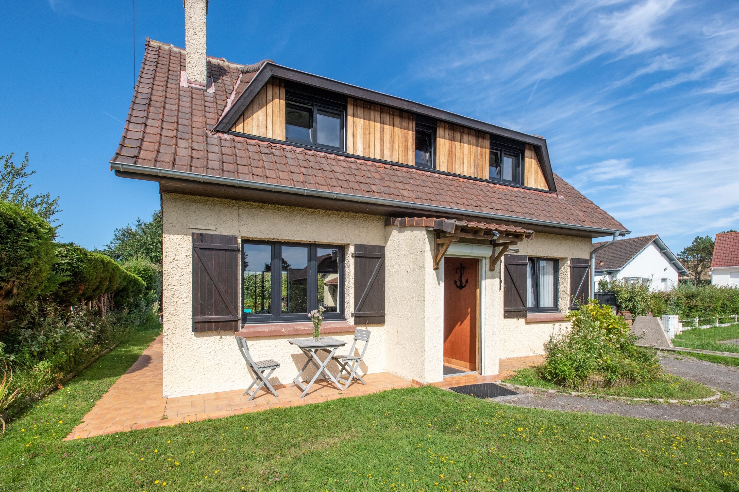 A two-story house with a tiled roof, a small front porch with a table and chairs, and a garden with a pathway.