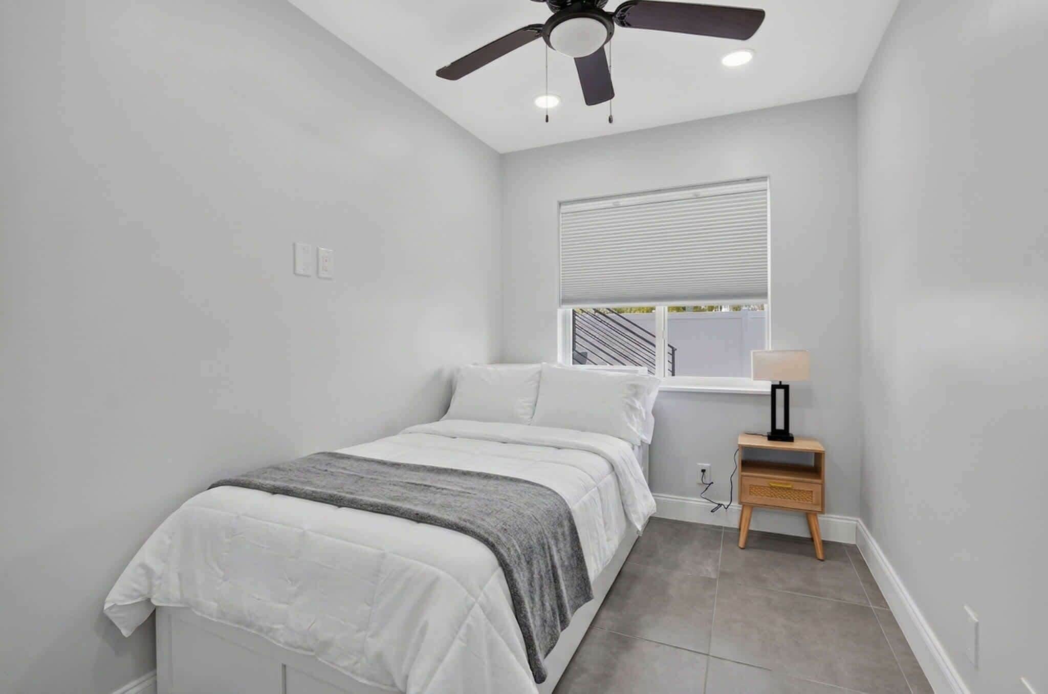 A bedroom with a bed, a ceiling fan, a window with blinds, a bedside table with a lamp, and a wall socket.