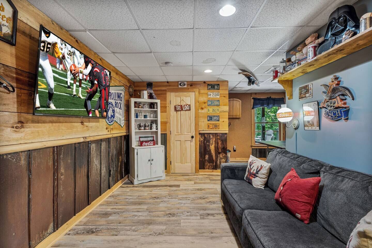 A room with a wooden wall, a television displaying a football game, a bookshelf, and a couch with pillows.