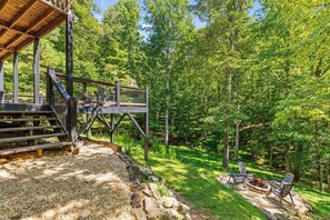 Une terrasse en bois avec un foyer et des chaises, entourée d’une zone boisée.
