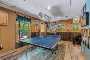 Une table de ping-pong dans une pièce aux murs en bois, une fenêtre avec des rideaux bleus et divers objets de collection liés au sport accrochés aux murs.