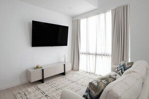 Ein modernes Wohnzimmer mit einem an der Wand befestigten Flachbildfernseher, einem weißen Sofa mit gemusterten Zierkissen, einem schlichten Konsolentisch und einem großen Fenster mit transparenten Vorhängen.