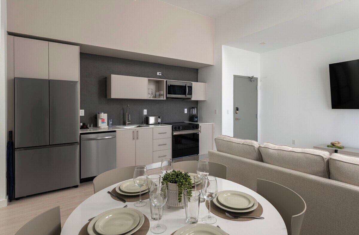 A modern kitchen with built-in appliances, a dining area with a round table set for four, and a living area with a sofa and flat-screen TV.