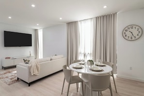 A modern living room with a sofa, dining table, and chairs. A flat-screen TV is mounted on the wall, and a clock is visible on the wall. - South Beach Apt: Rooftop Pool/Prime Location #201 (Miami Beach)