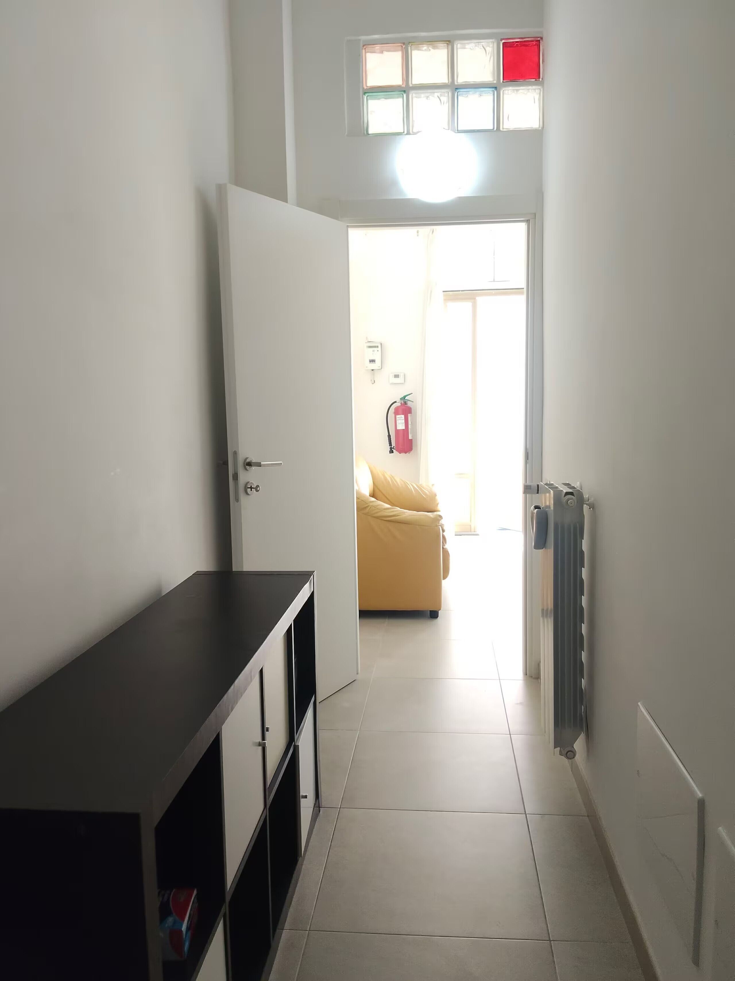 Un corridoio con una porta che dà su una stanza, un termosifone e un estintore a parete.
