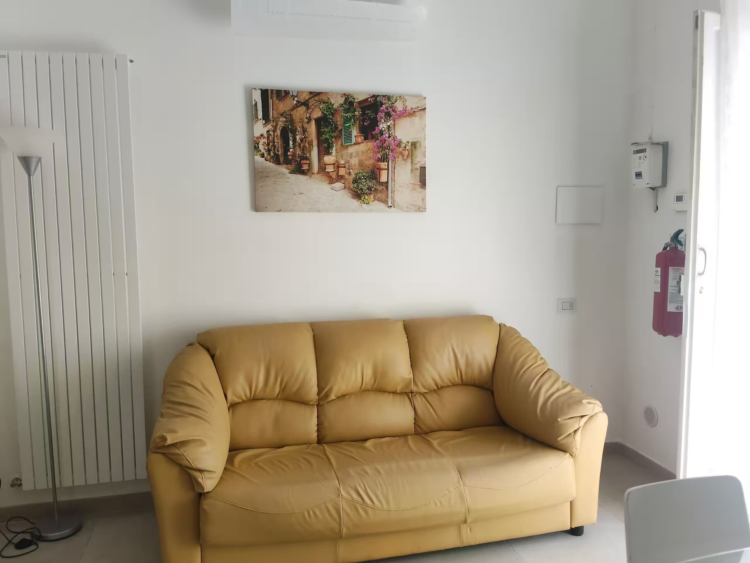 Ein Zimmer mit einem beigen Sofa, einem Heizkörper, einem Wandbild und einem Feuerlöscher.