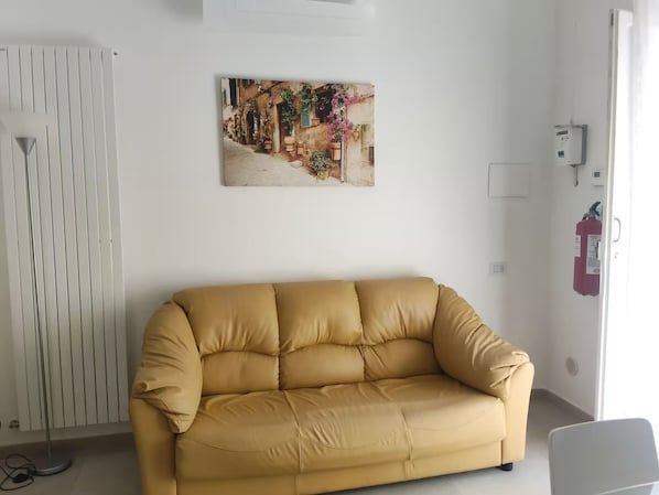 A room with a beige sofa, a radiator, a wall-mounted picture, and a fire extinguisher.