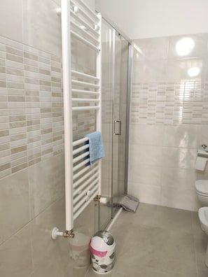 A bathroom with a glass shower enclosure, a white towel rack with a blue towel, a toilet, and a trash can.