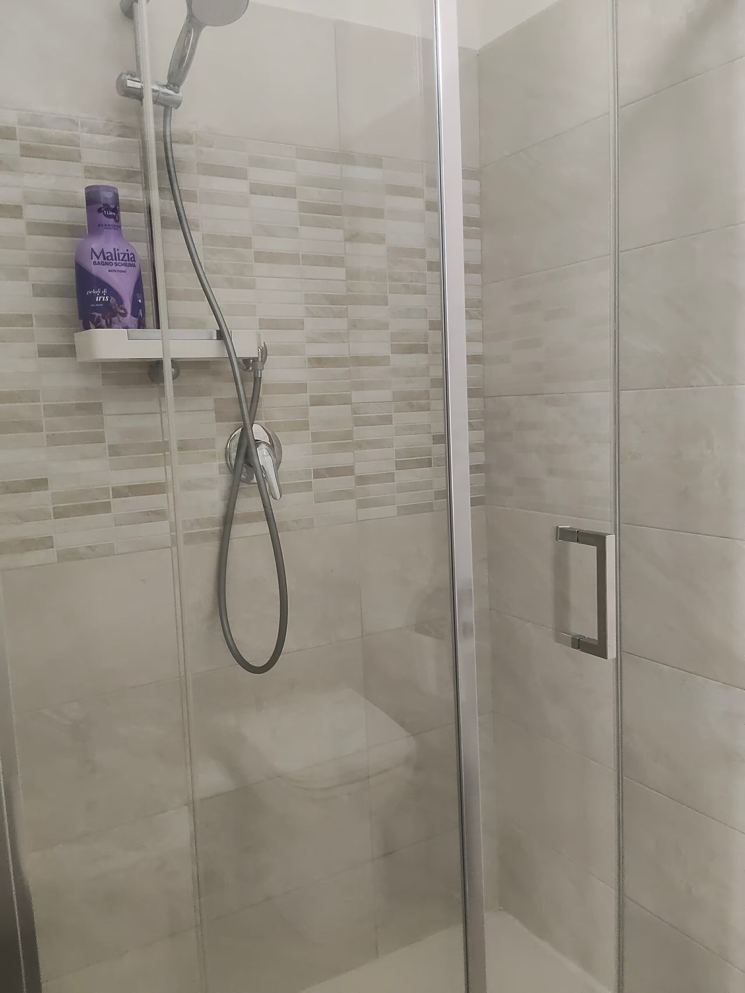 Un espace douche doté d’une porte en verre, d’un pommeau de douche à main et d’une étagère sur laquelle se trouve une bouteille.