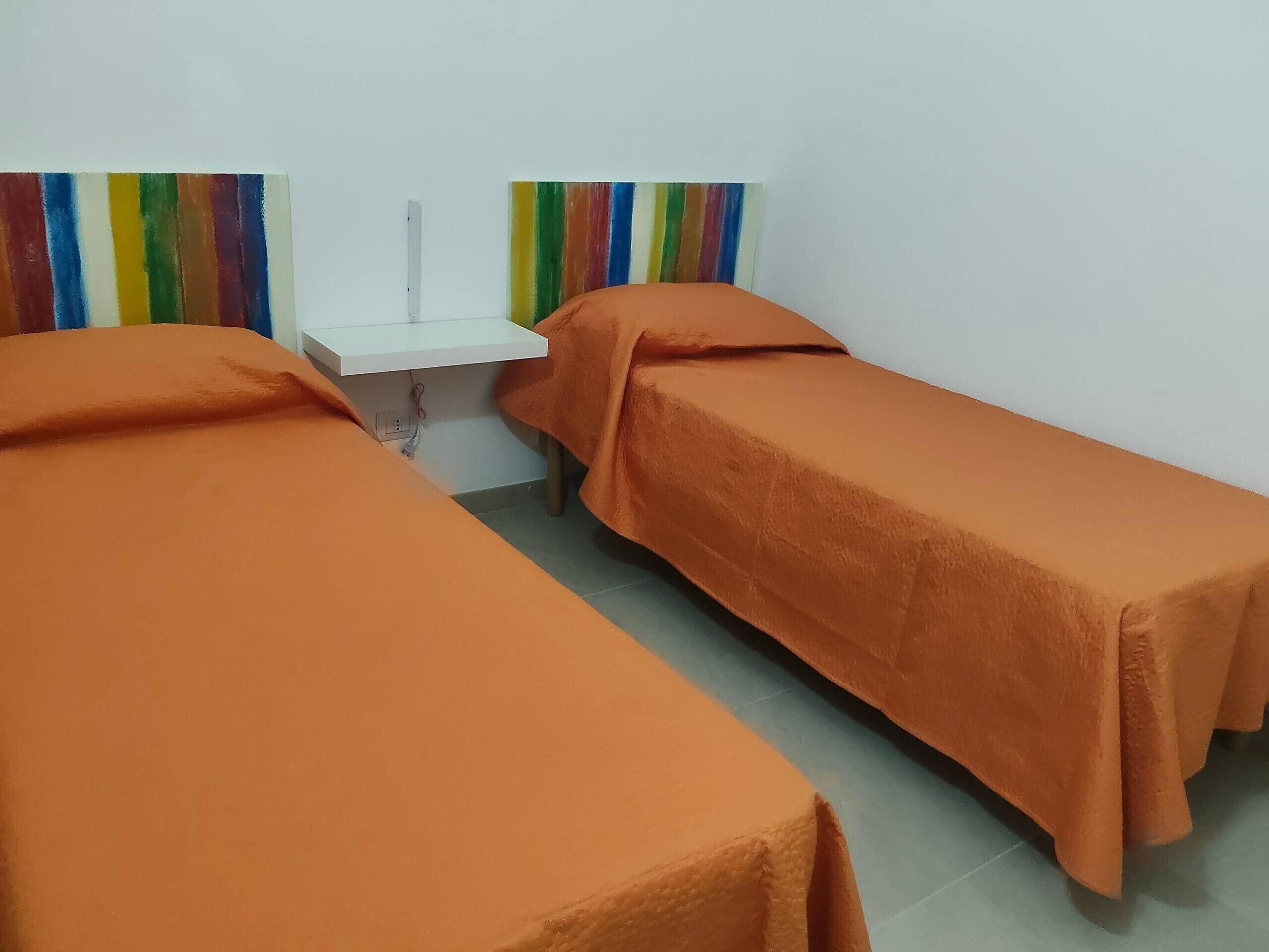 Ein Zimmer mit zwei Betten, jedes mit einer orangefarbenen Decke, einem weißen Nachttisch und einer farbenfrohen Wanddekoration mit Streifen.