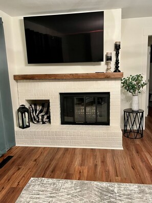 A living room with a fireplace, a flat-screen TV mounted above it, and a potted plant on a side table.