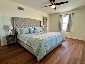 A bedroom with a tufted headboard, a bed with light blue bedding, a ceiling fan, a nightstand with a lamp, and a window with curtains.