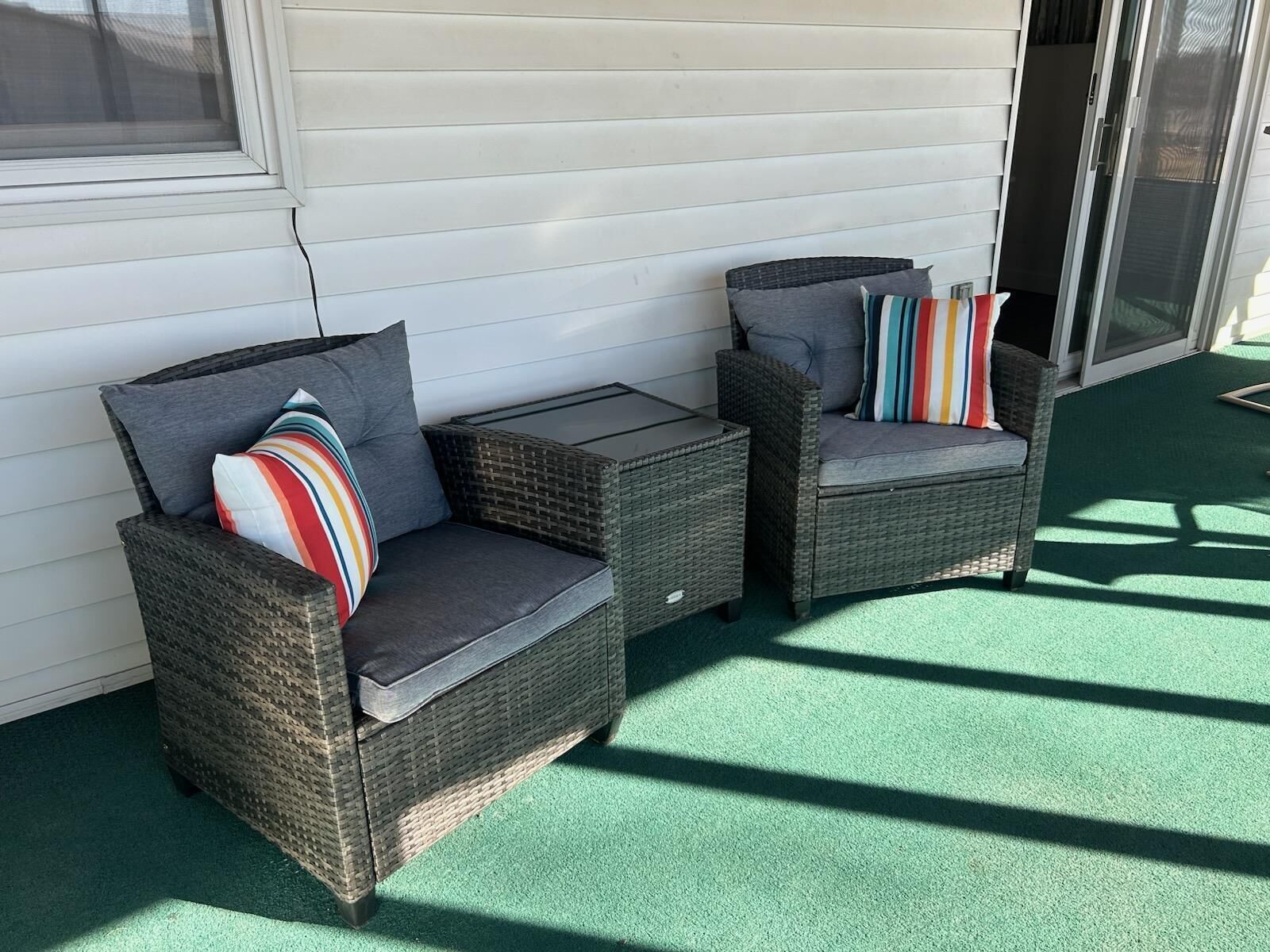 Two wicker chairs with cushions and a small table, positioned on a green surface with a white wall and a sliding glass door in the background.