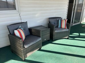 Two wicker chairs with cushions and a small table, positioned on a green surface with a white wall and a sliding glass door in the background.