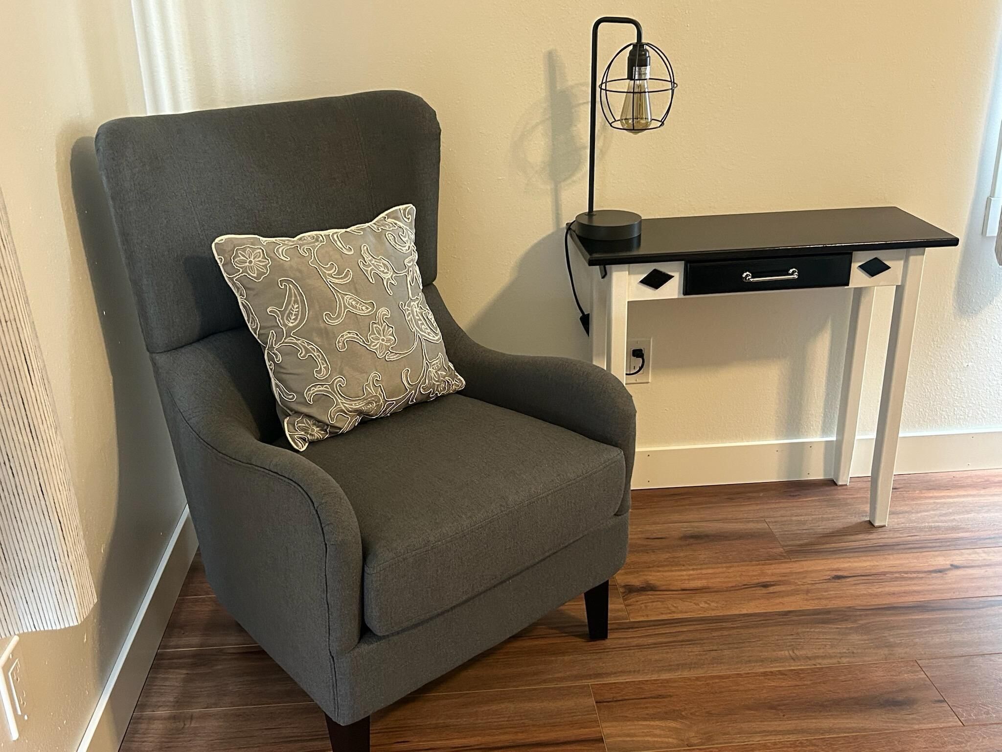 A grey armchair with a patterned pillow, a black desk with drawers, and a wall-mounted lamp.