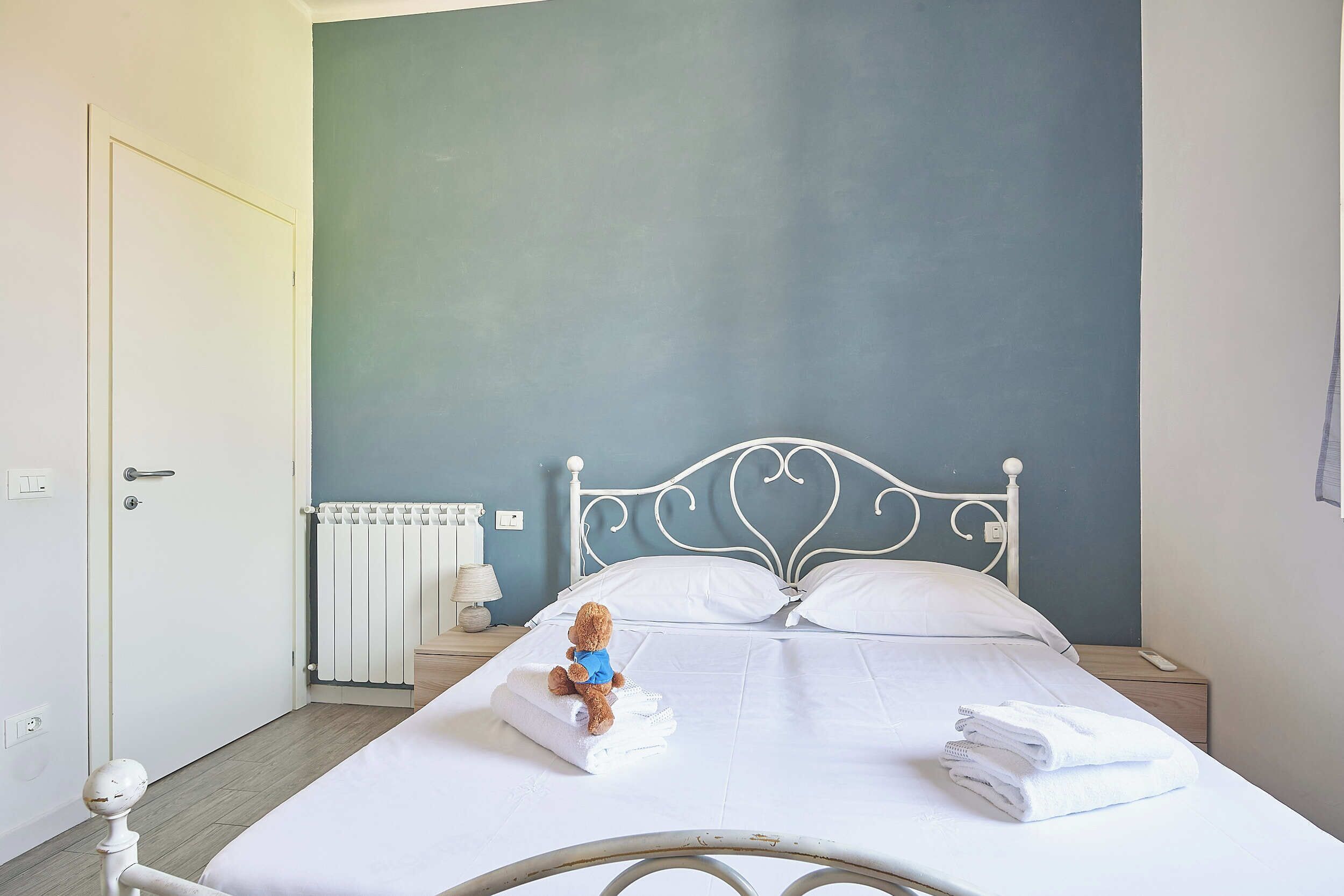 A bedroom with a bed, a nightstand, a radiator, and a teddy bear on the bed.