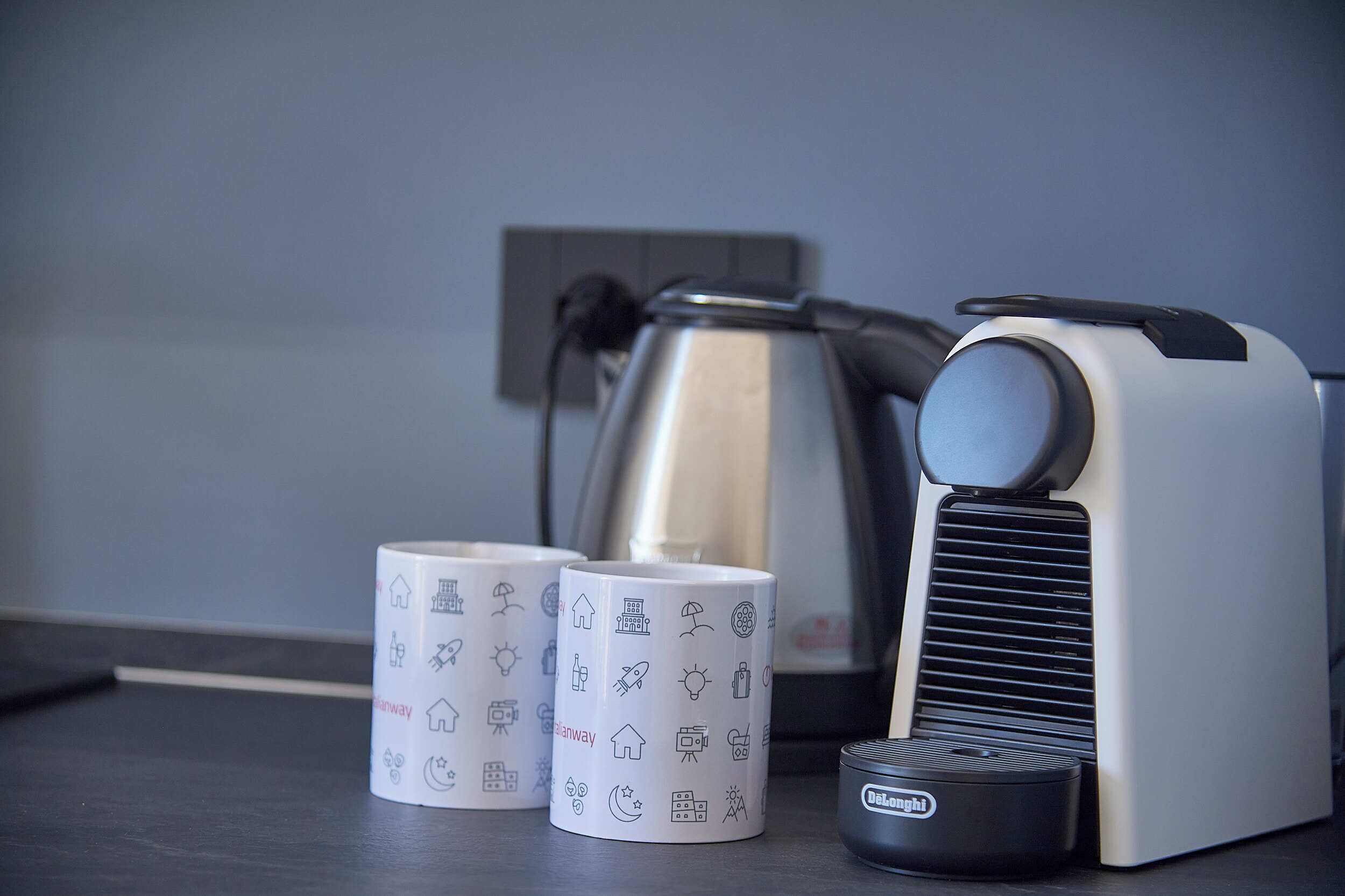 A DeLonghi coffee maker, two mugs with travel-themed designs, and a kettle on a kitchen counter.