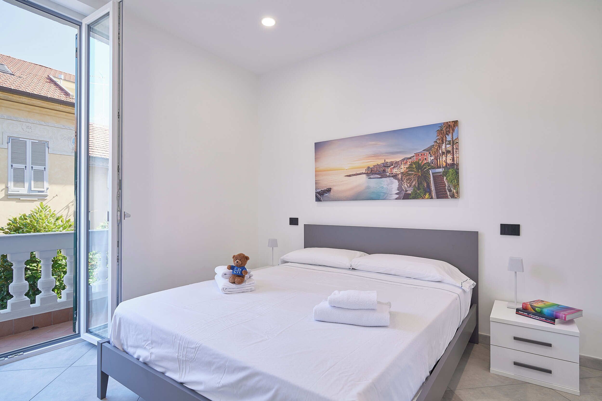 Una habitación de hotel con una cama, una mesita de noche, una lámpara, un osito de peluche y un cuadro de un pueblo costero.