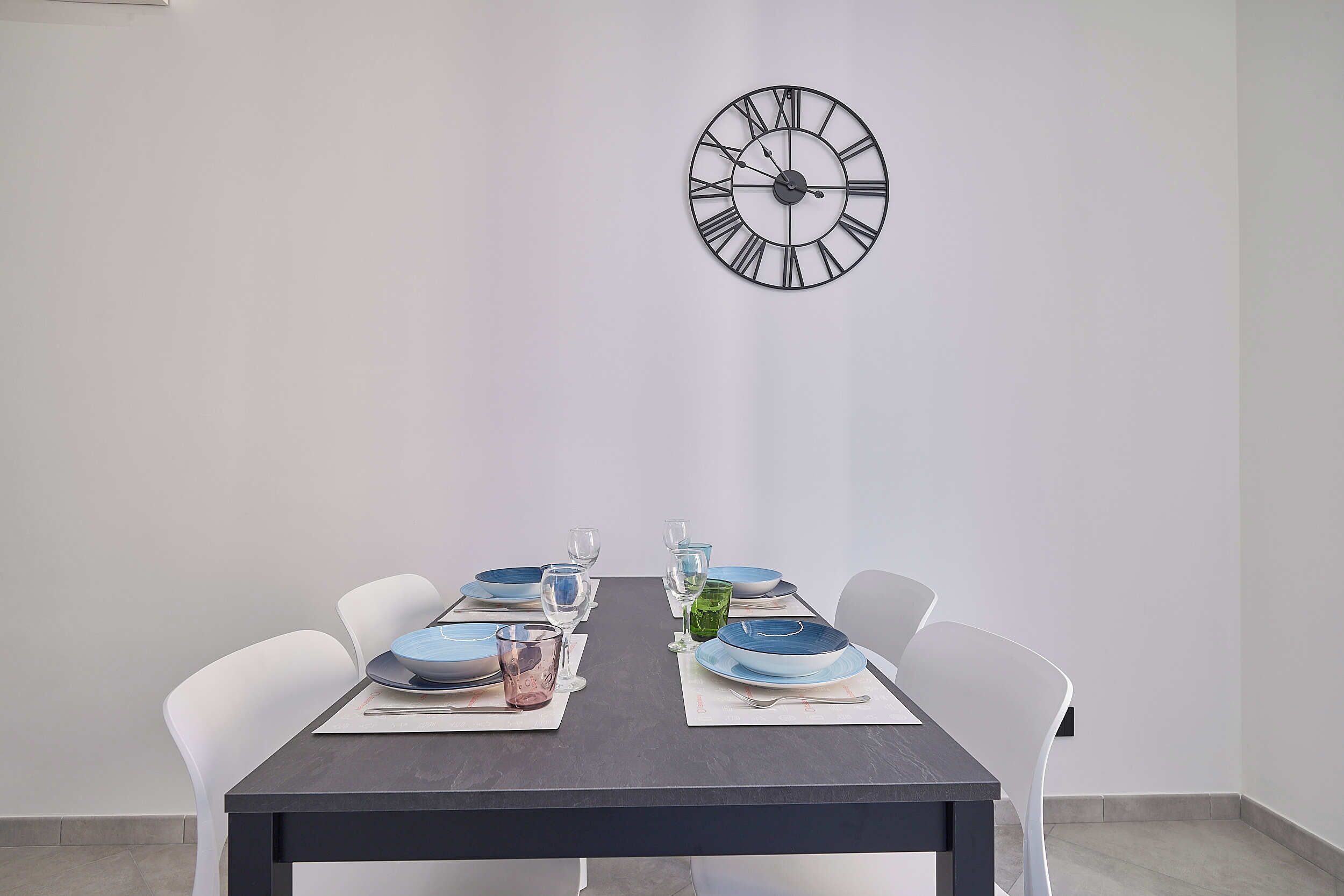 Une table à manger avec une horloge au mur, des chaises blanches et des assiettes bleues et vertes.
