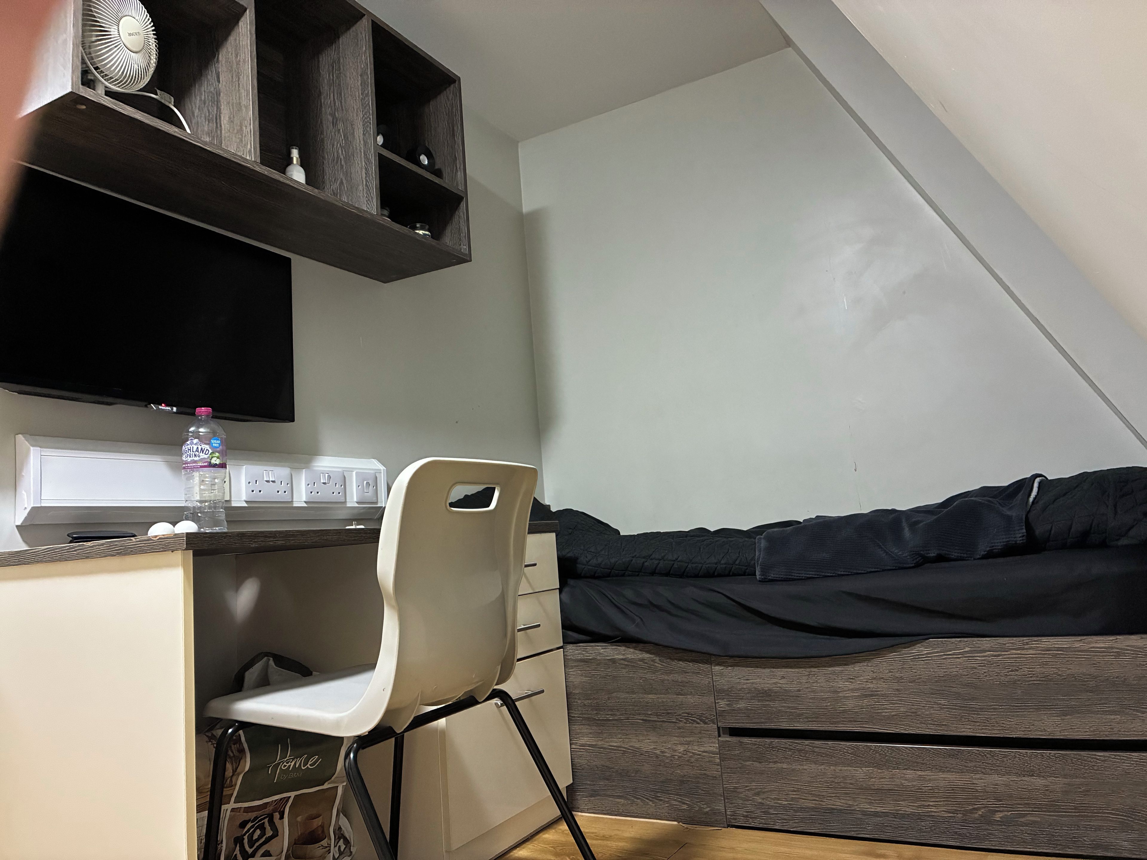 A small room with a desk, chair, and a bed with a dark cover. There is a water bottle on the desk and a power strip with outlets.