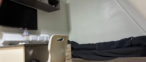 A small room with a desk, chair, and a bed with a dark cover. There is a water bottle on the desk and a power strip with outlets.