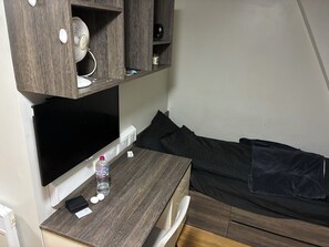 Habitación pequeña con una televisión de pantalla plana montada en una pared de madera, una mesita de madera con una botella de agua, un control remoto y una billetera. Incluye una cama con una manta oscura y almohadas.