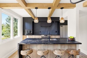 A modern kitchen with a black marble island, wooden stools, and pendant lighting. - Renovated Alpine Retreat | Ski & Golf Views (The Blue Mountains)