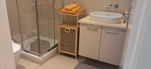 A modern bathroom with a glass shower enclosure, a wooden shelf with towels, a sink, and a toilet. - Vrbo Property (Luanda)