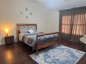 Ein Schlafzimmer mit einem hölzernen Bett, einem Nachttisch, einem blauen Teppich mit Muster und einem Fenster mit Vorhängen.